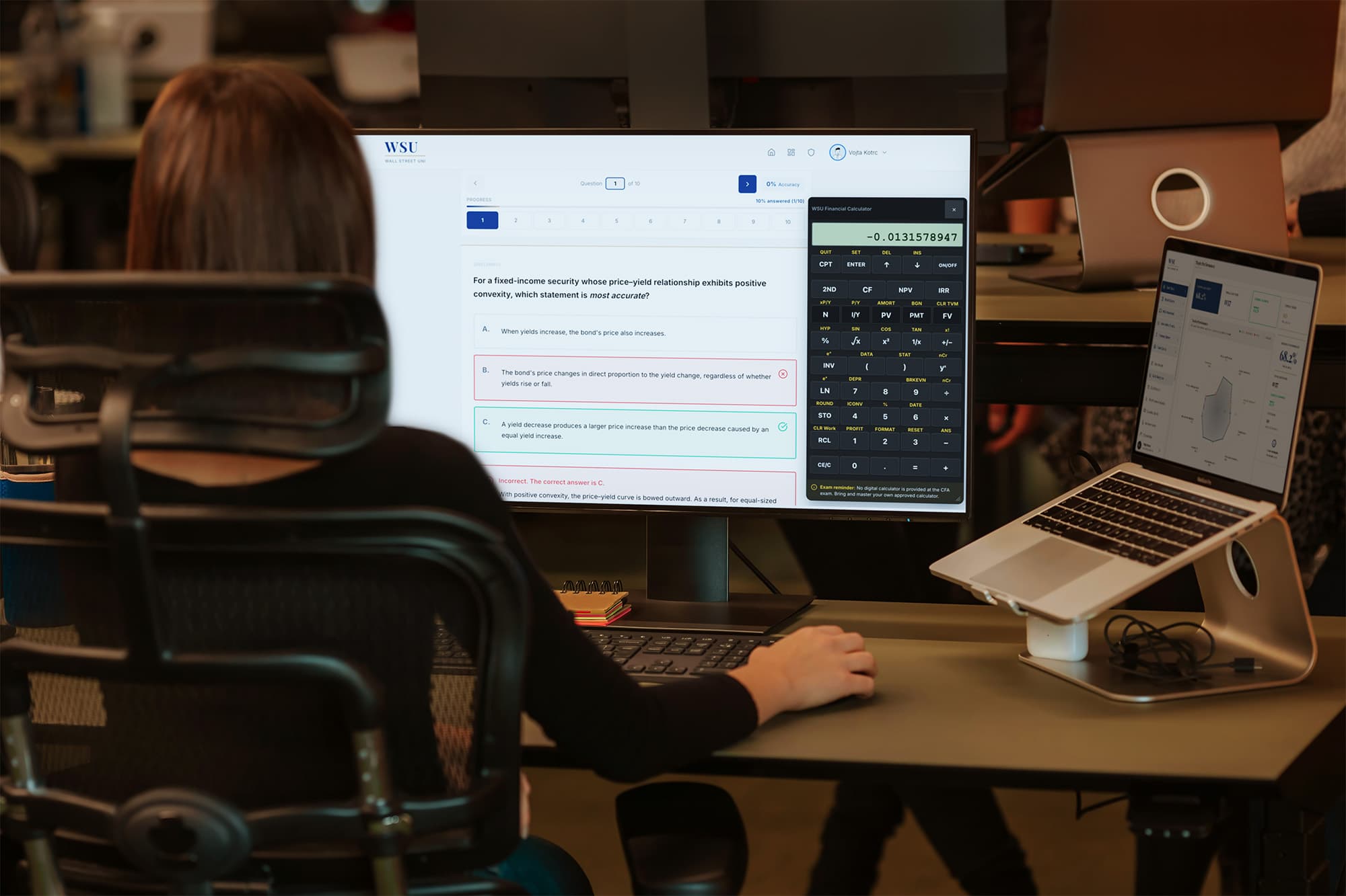 A candidate at a desk working through a fixed-income convexity question on Wall Street Uni, with the financial calculator open on the monitor and topic analytics on a laptop beside it